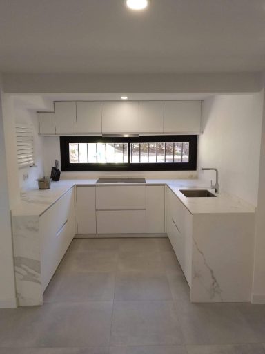 Cocina moderna con armarios blancos y ventana grande, suelo de baldosas grises.