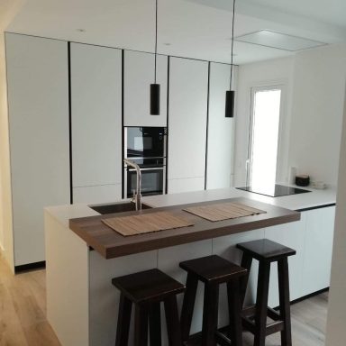 Cocina moderna con armarios blancos, barra de madera y taburetes oscuros.