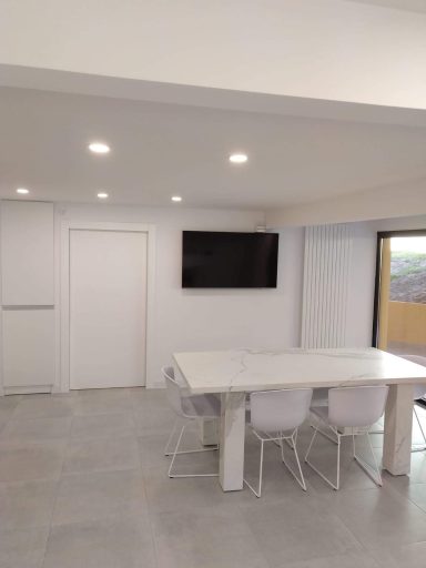 Interior de una sala moderna con mesa blanca, sillas transparentes y televisor en la pared.
